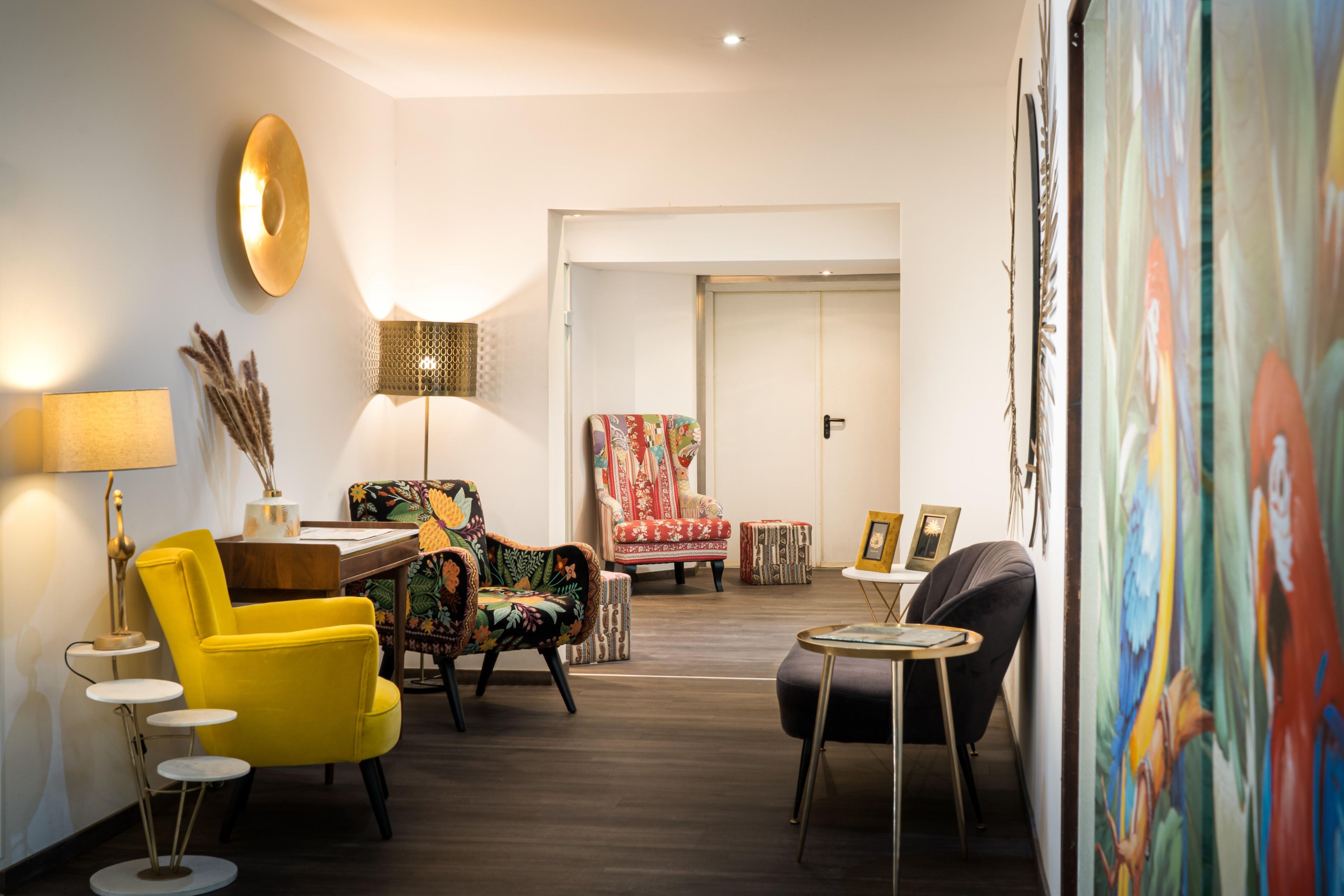 A modern living room with a yellow armchair, a patterned armchair, a red patterned chair, and a black armchair. There is a small round table with a lamp and a decorative vase.