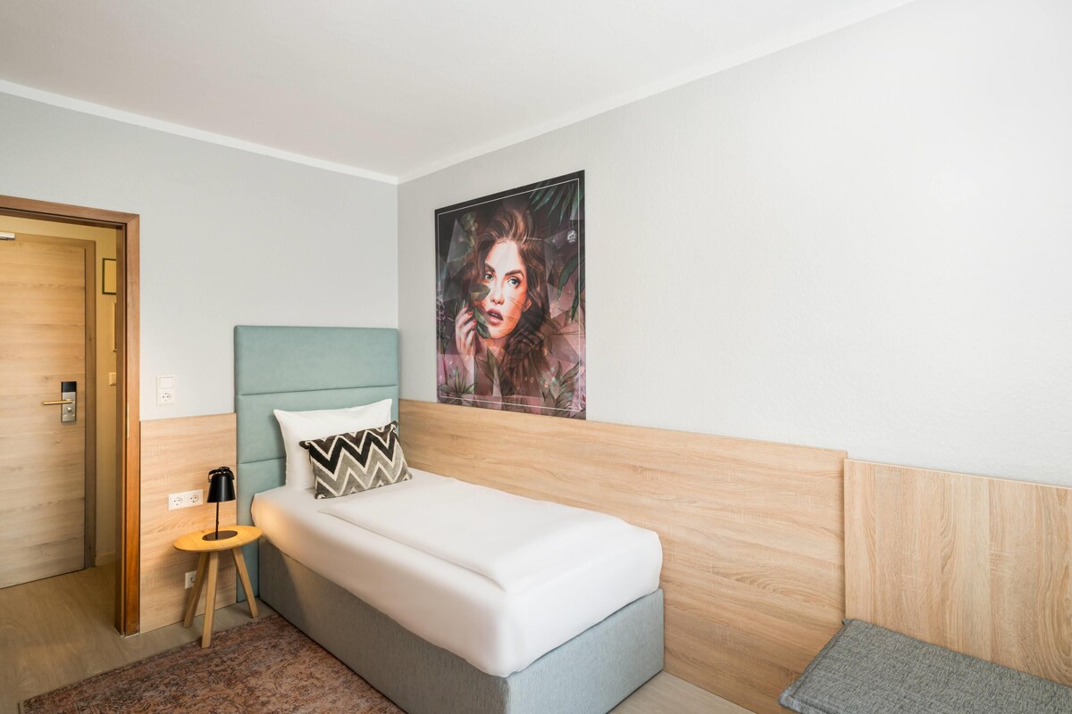 A bedroom with a bed, a nightstand, a wall-mounted lamp, a door, and a poster on the wall.