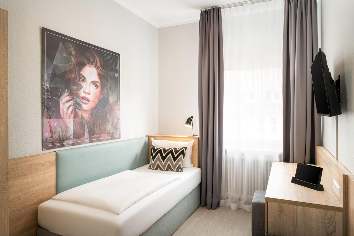 A modern hotel room with a bed, a desk with a tablet, a painting on the wall, and a window with curtains.