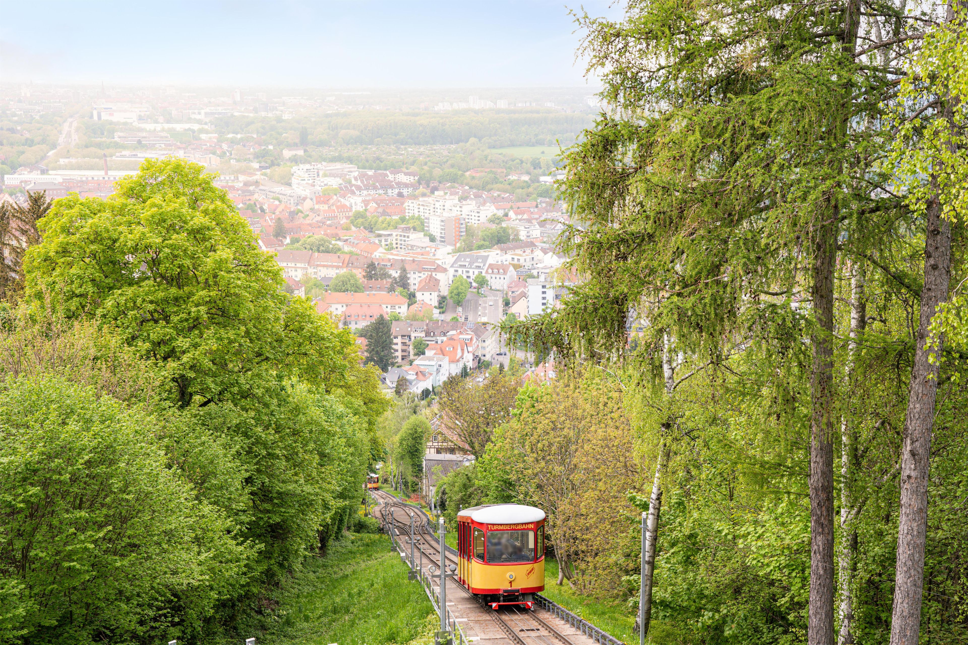 A yellow train on a track surrounded by lush greenery and a cityscape in the background.