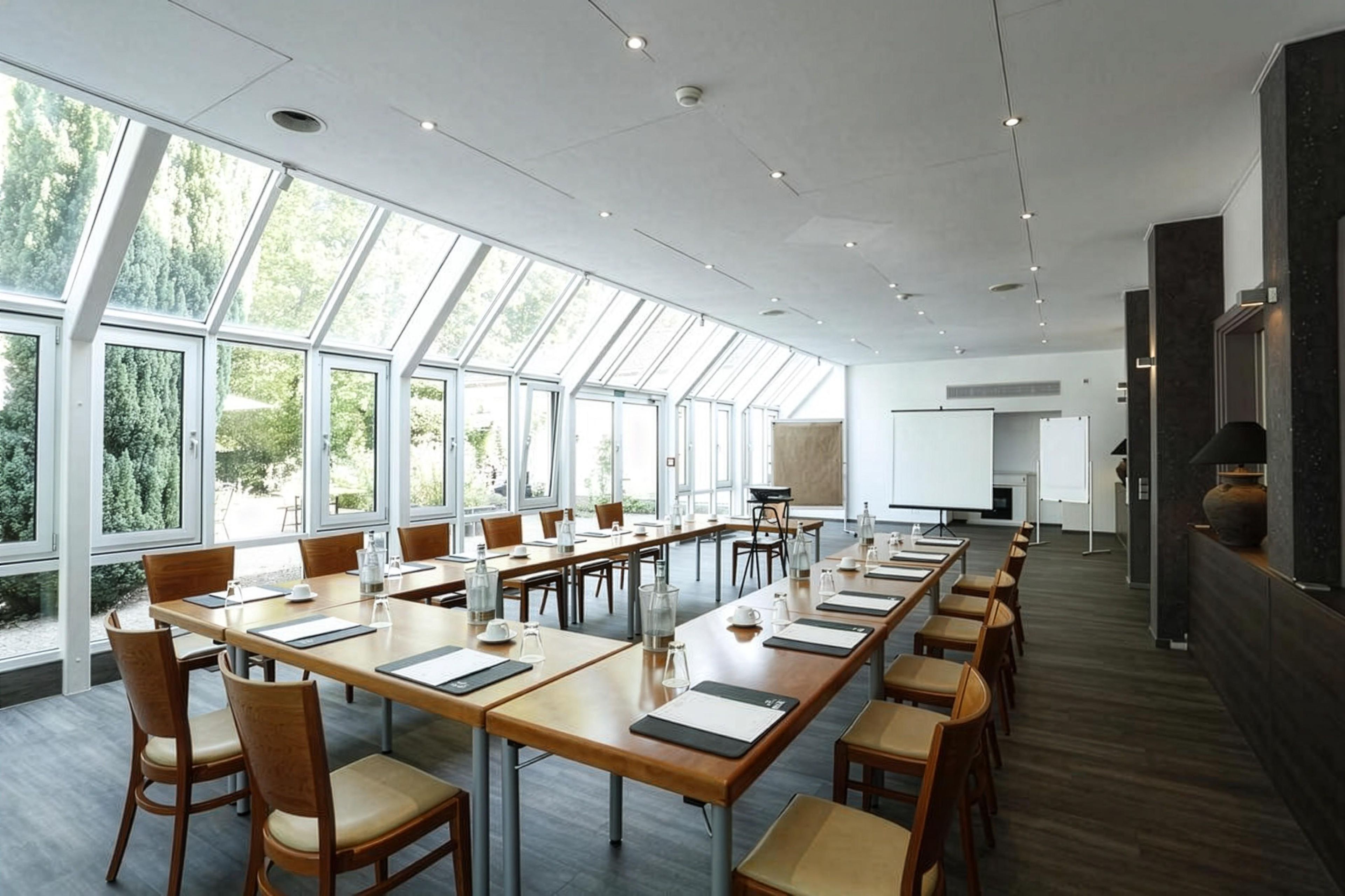 Ein Konferenzraum mit langen Tischen und Stühlen, großen Fenstern und einer Whiteboard-Tafel.
