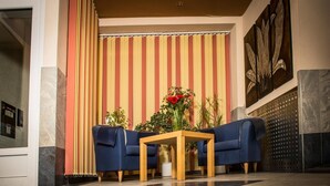 A lobby with two blue armchairs, a wooden coffee table, and a striped wall with a large plant and flowers. - Hotel Aria (Frankfurt)