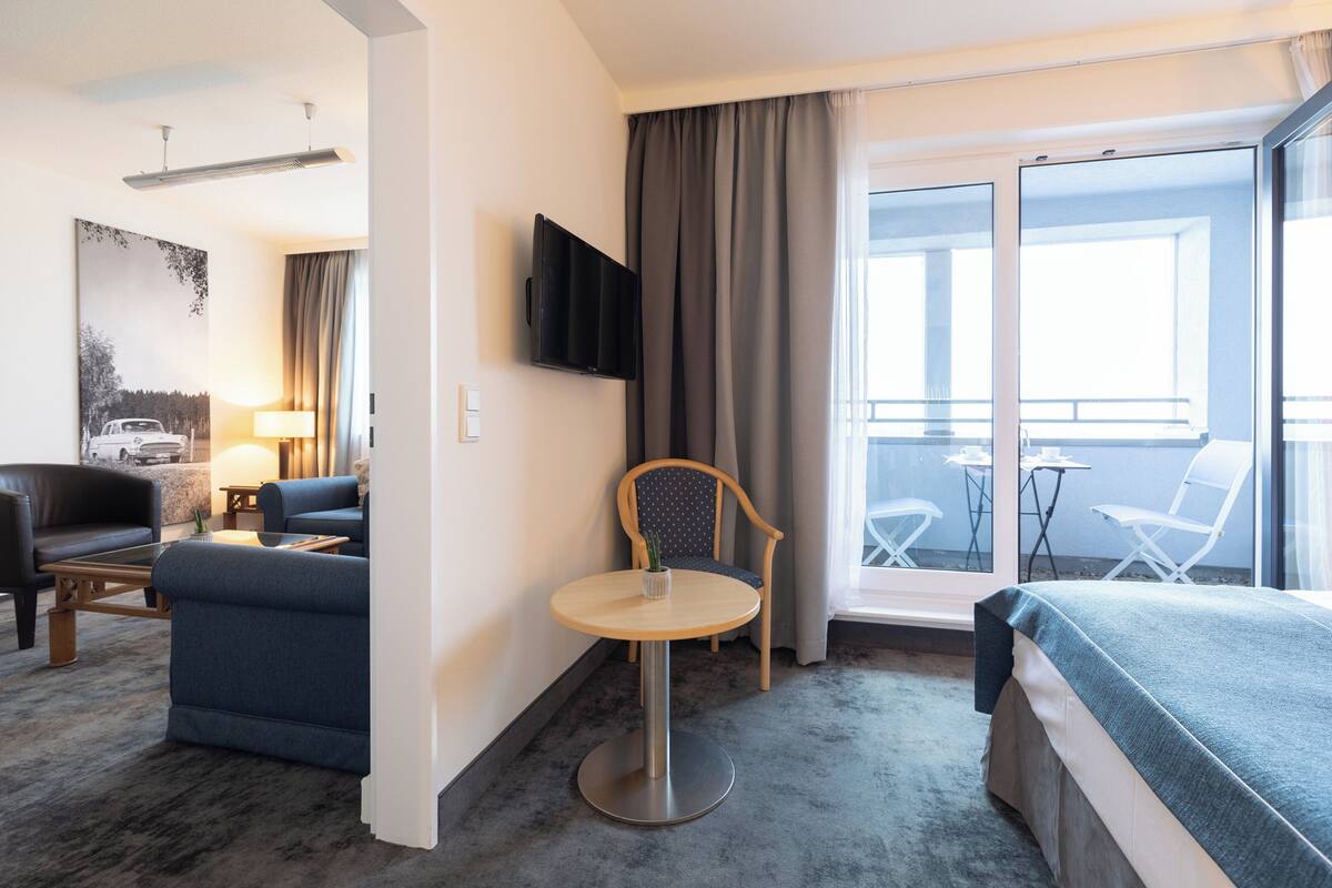 A hotel room with a bed, a round table, a sofa, a chair, a TV, and a balcony with chairs.