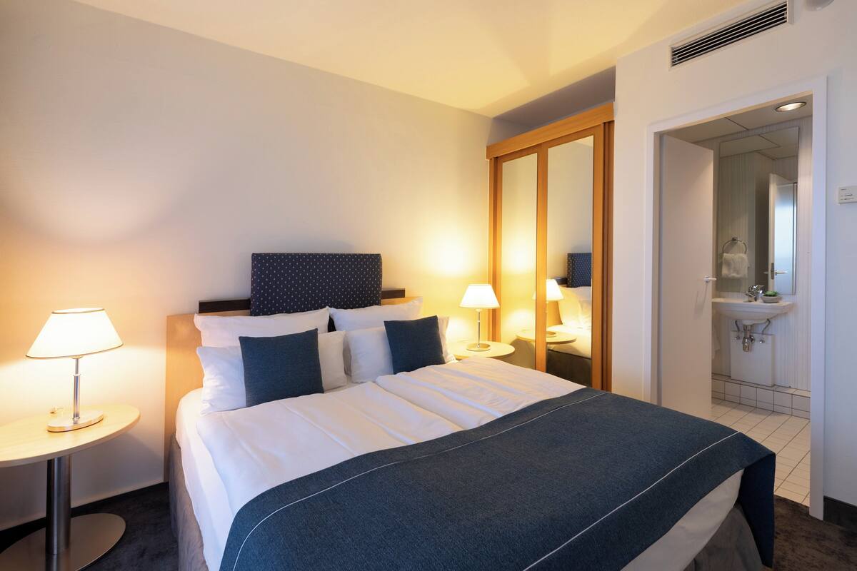 A hotel room with a neatly made bed, two bedside lamps, a round table, and a bathroom visible through an open door.