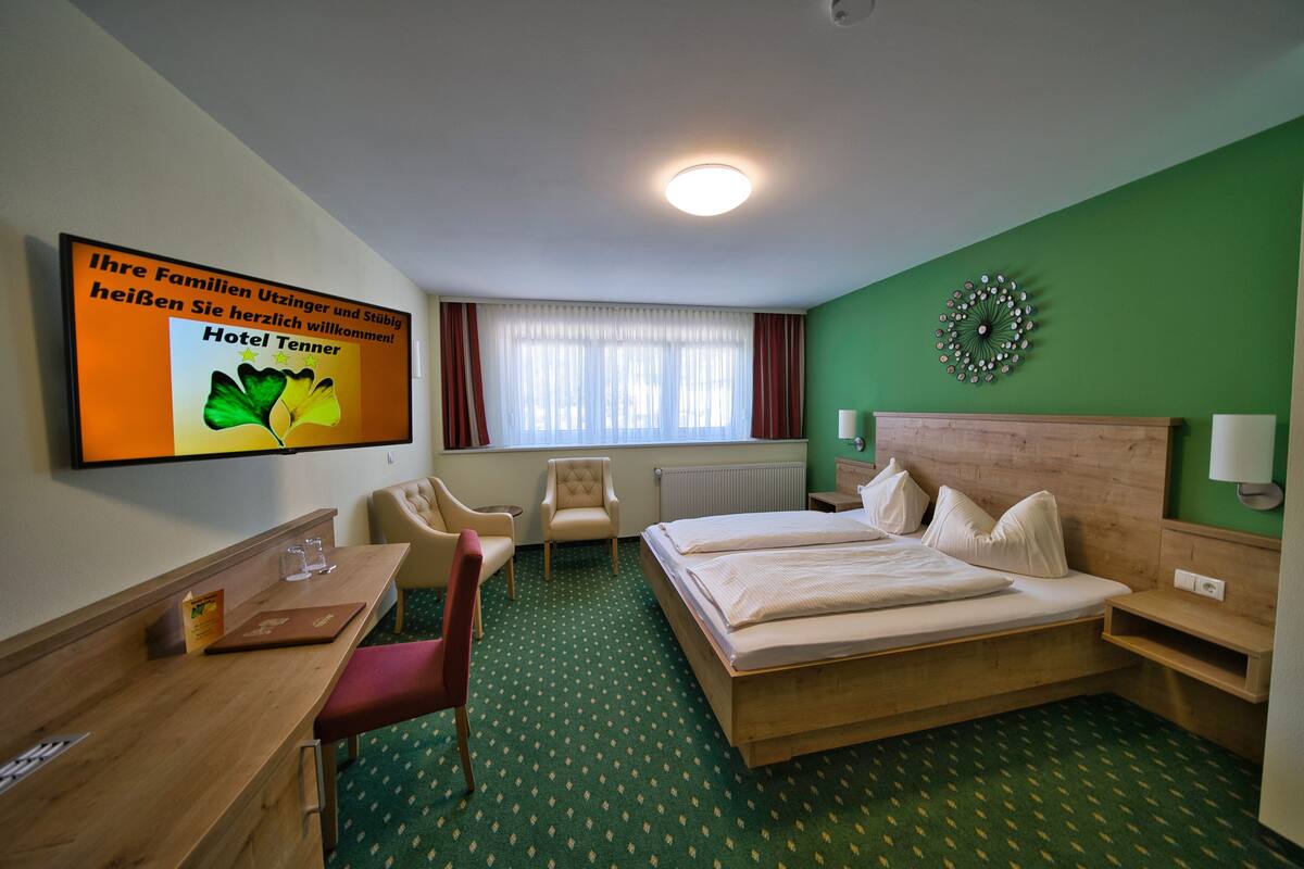 Quarto de hotel com cama, escrivaninha, cadeira, mesa pequena e televisão exibindo uma mensagem de boas-vindas.