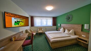 Quarto de hotel com cama, escrivaninha, cadeira, mesa pequena e televisão exibindo uma mensagem de boas-vindas.
