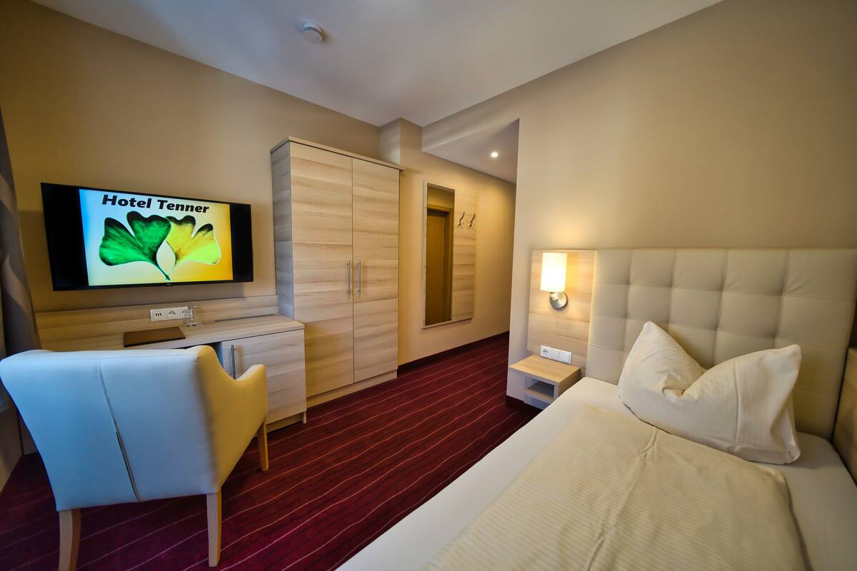 Quarto de hotel com cama, escrivaninha, cadeira, TV exibindo 'Hotel Tenner' e armário.