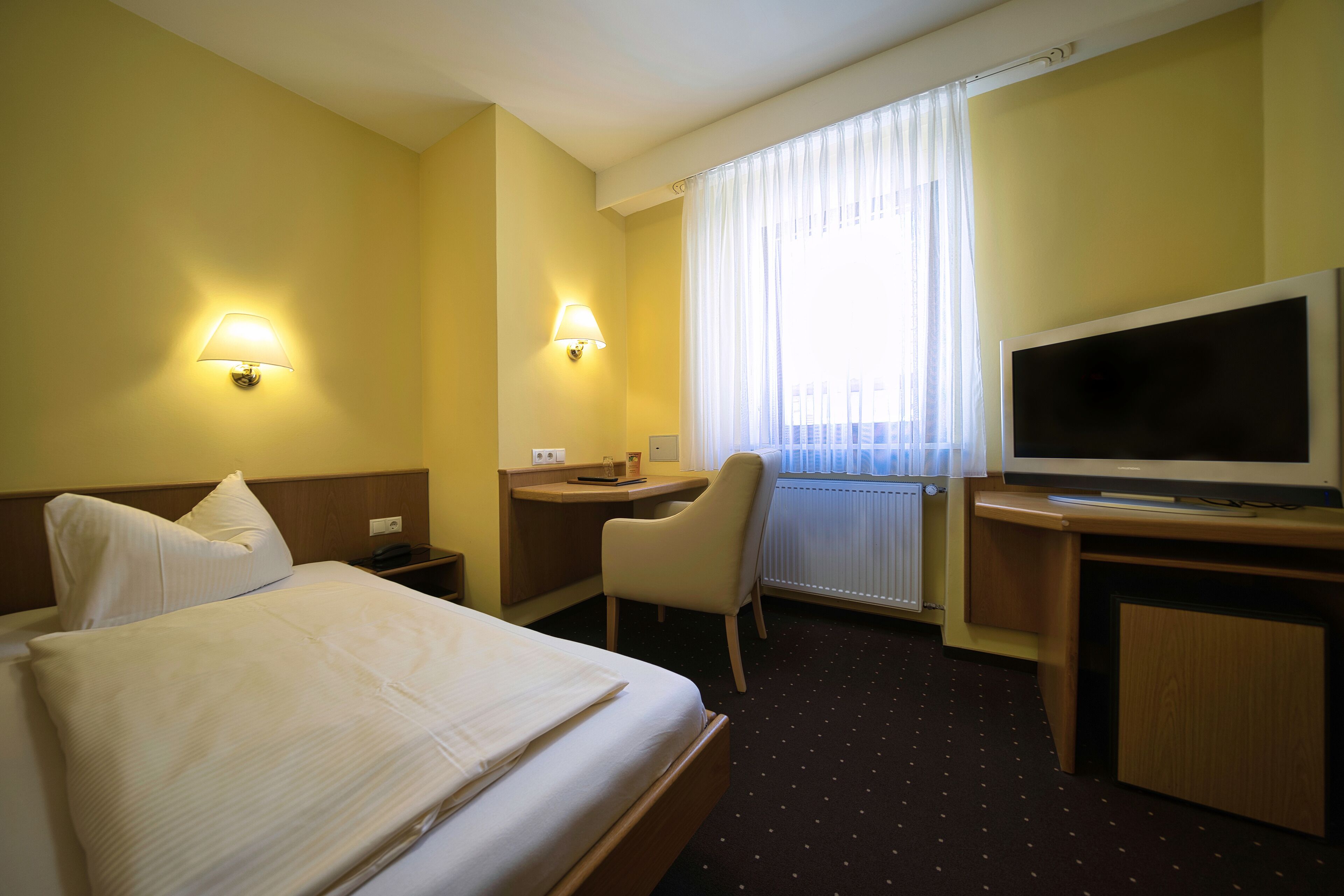 Una habitación de hotel con cama, escritorio, silla, televisor y ventana con cortinas.