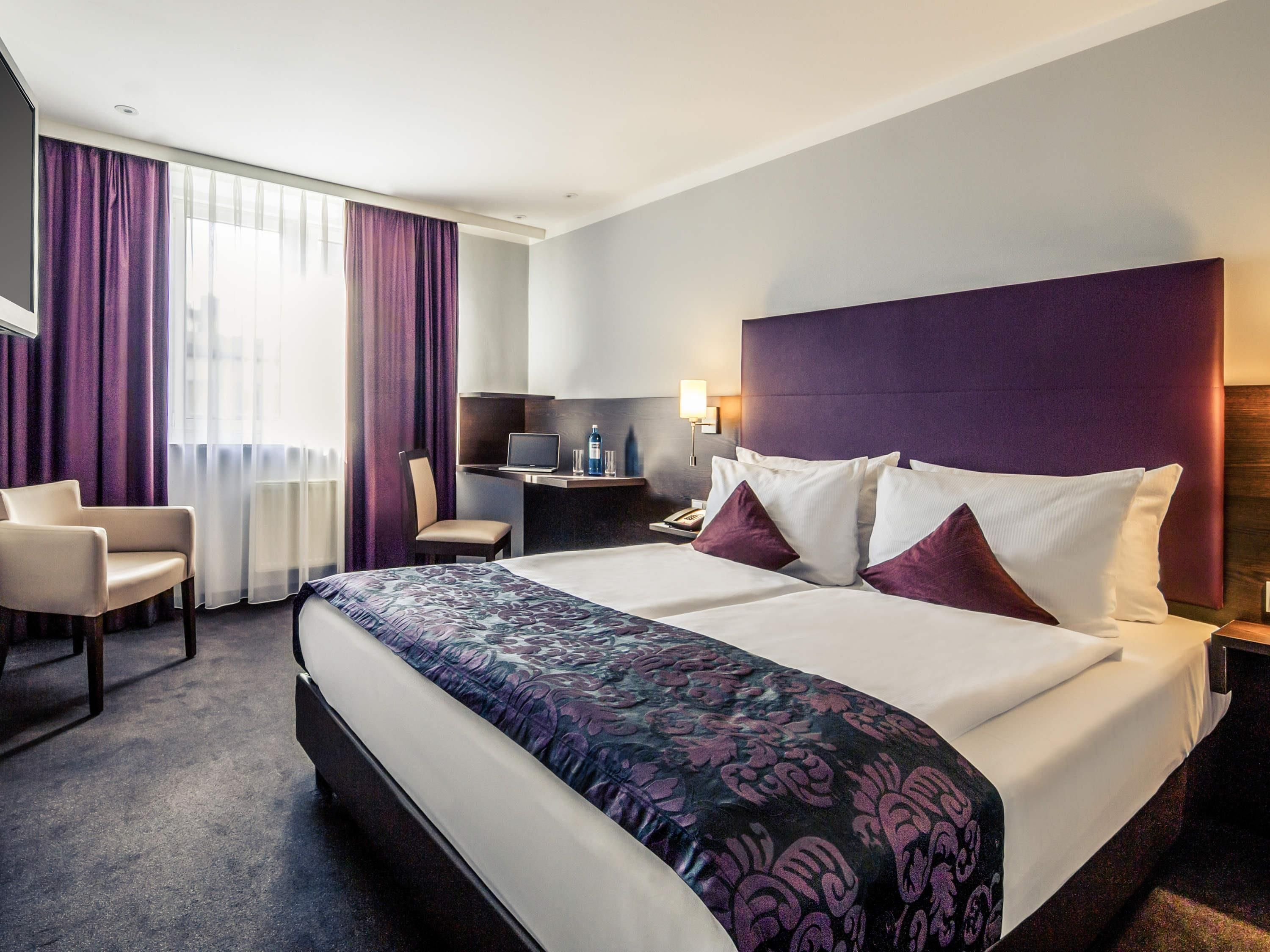 A hotel room with a large bed, a desk, a chair, and a view of the city through the window.