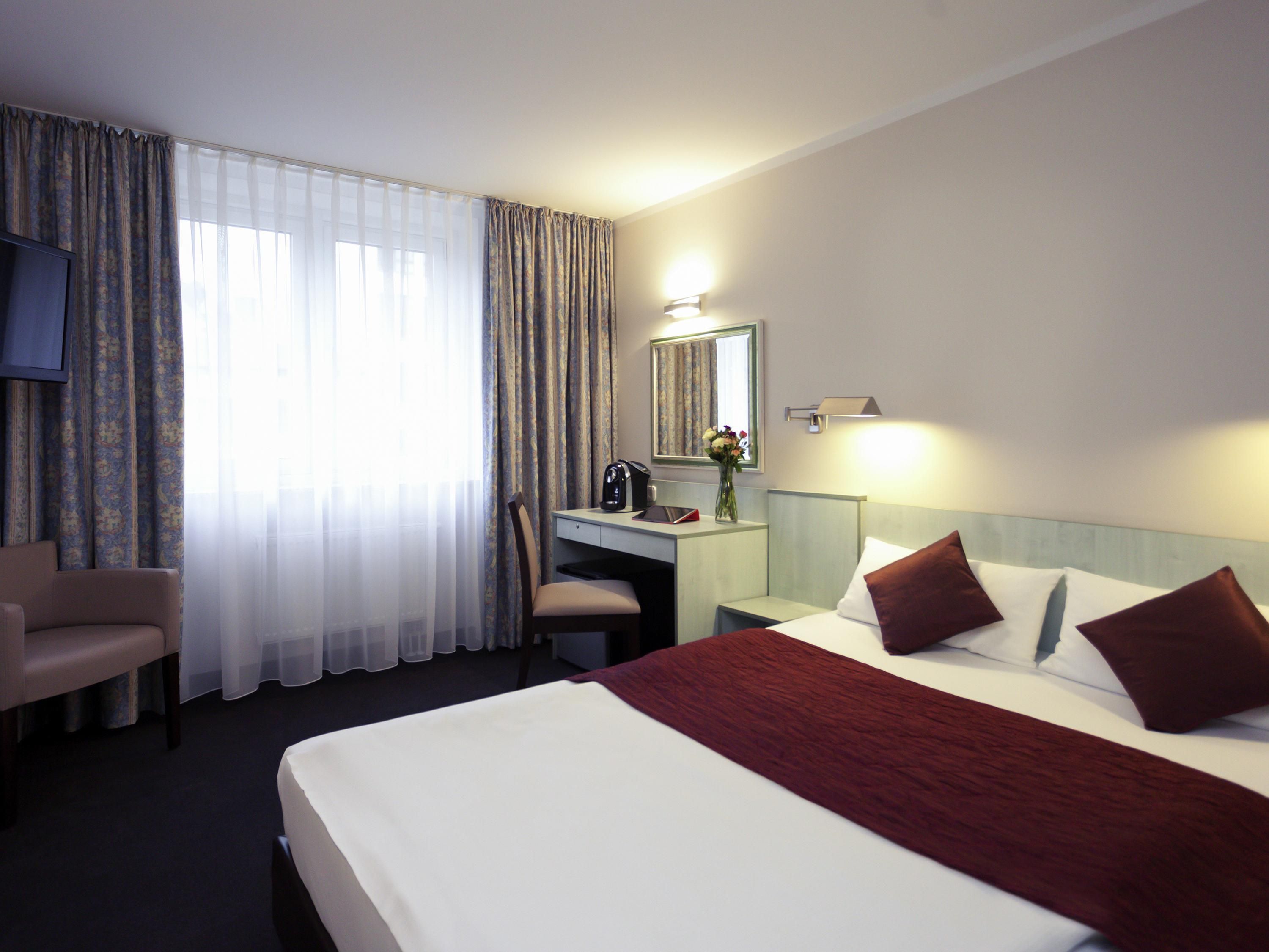 Habitación de hotel con cama, escritorio, silla, televisor y ventana con cortinas.