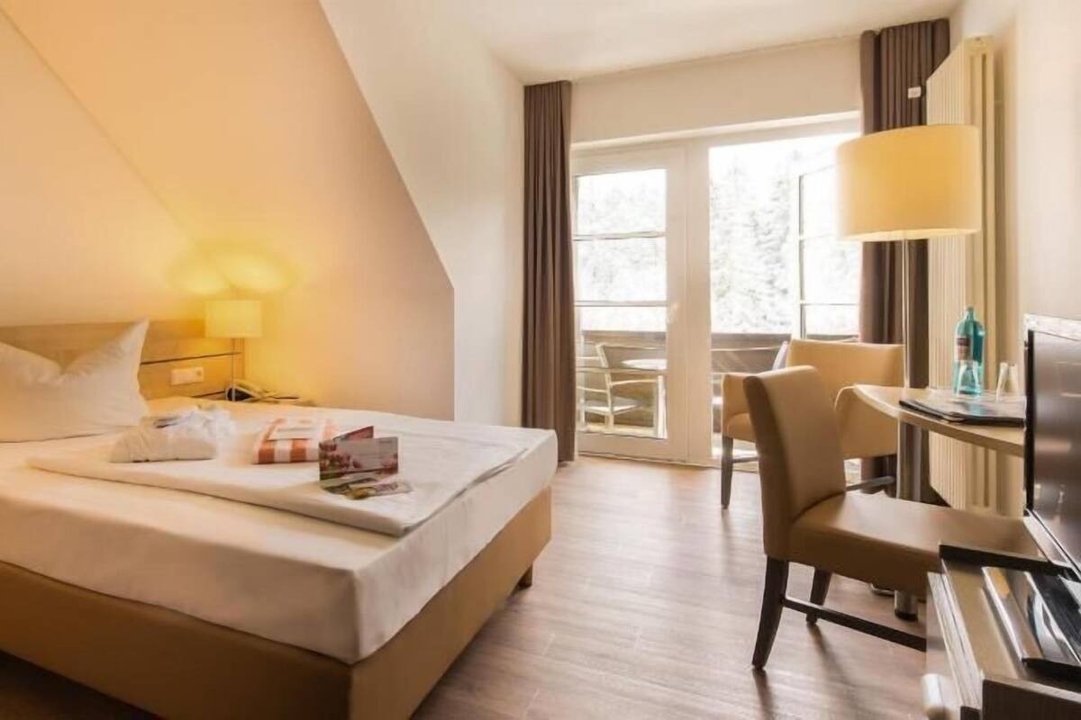 Ein Hotelzimmer mit einem Bett, Nachttischen, einem Schreibtisch mit Stuhl, einer Lampe und Blick ins Freie.