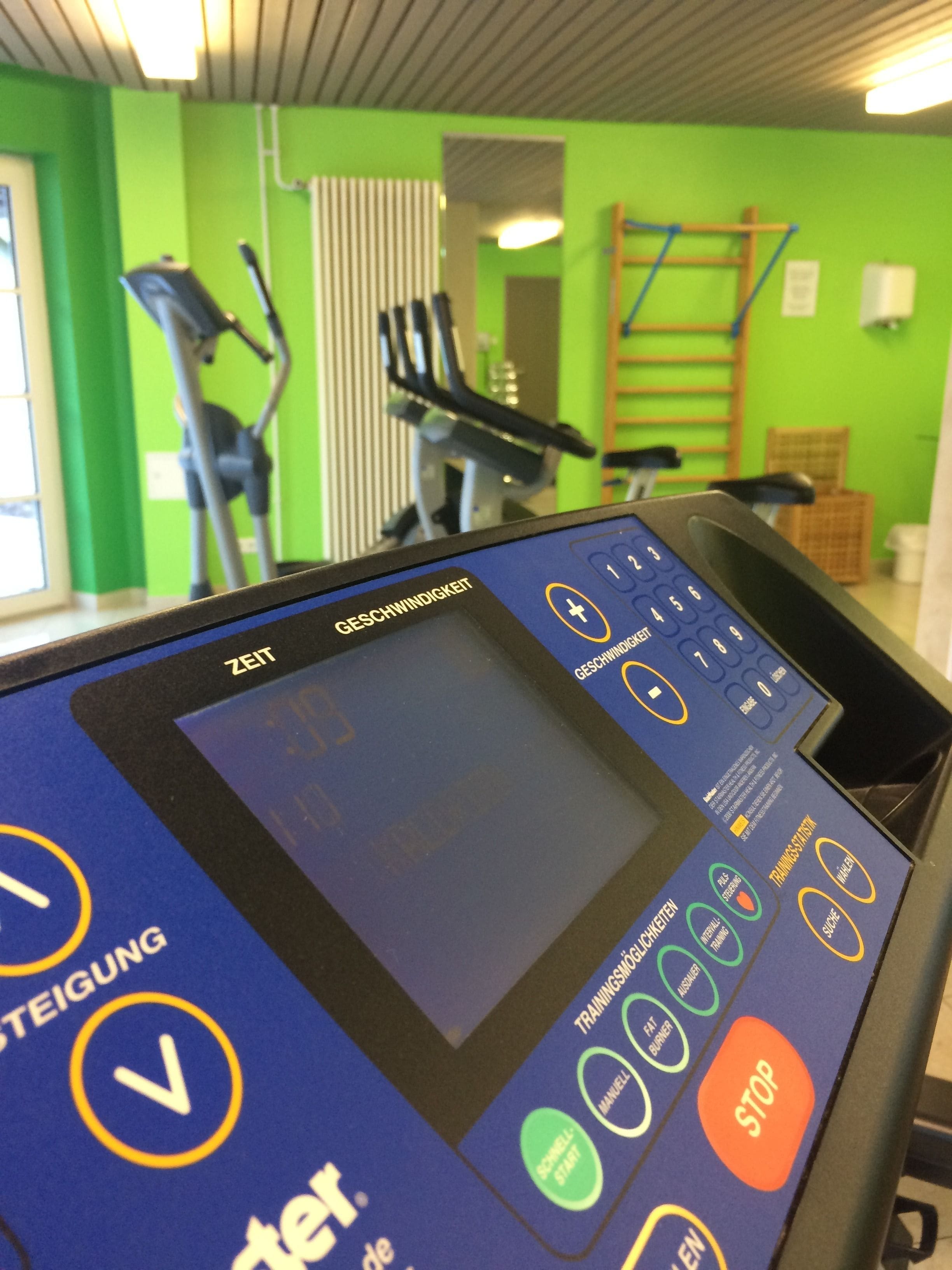A fitness machine with a digital display and control panel, surrounded by exercise equipment and a green wall.