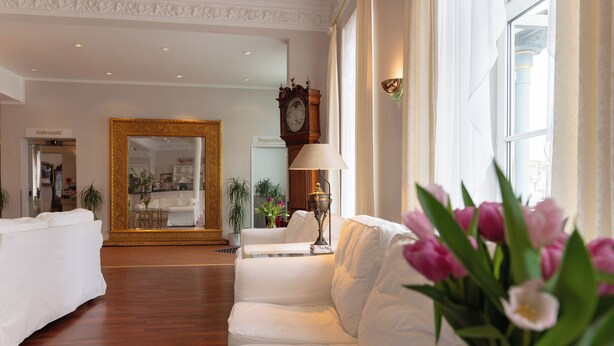 Un vestíbulo de hotel con un sofá blanco, un espejo, un reloj y un jarrón de flores.