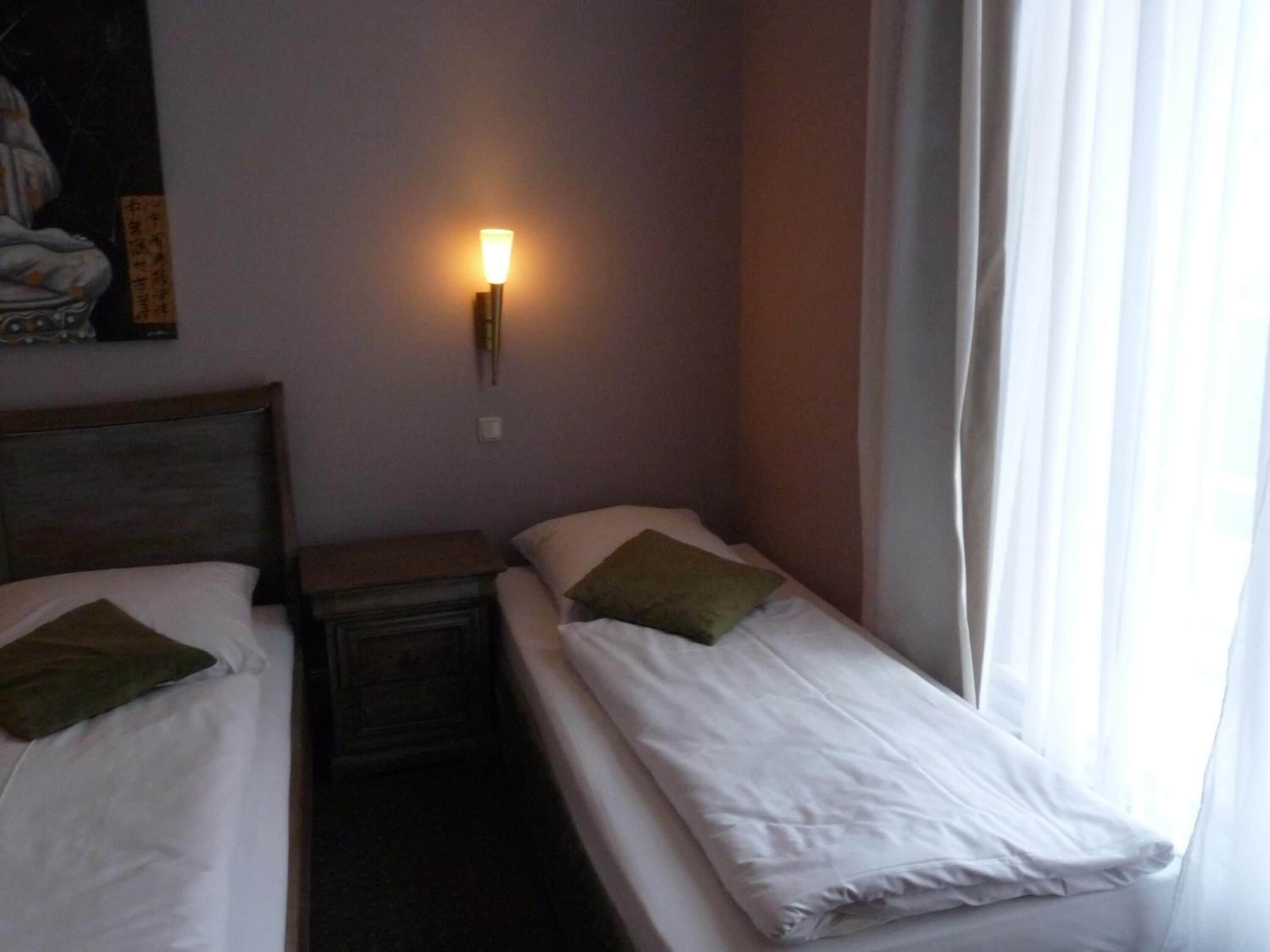 A hotel room with two single beds, a nightstand, a wall-mounted lamp, and a window with curtains.