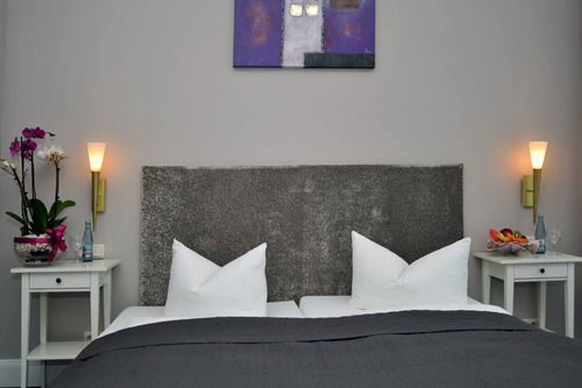Ein ordentlich bezogenes Bett mit weißen Kissen und einer dunklen Tagesdecke, begleitet von zwei Nachttischen mit Lampen.