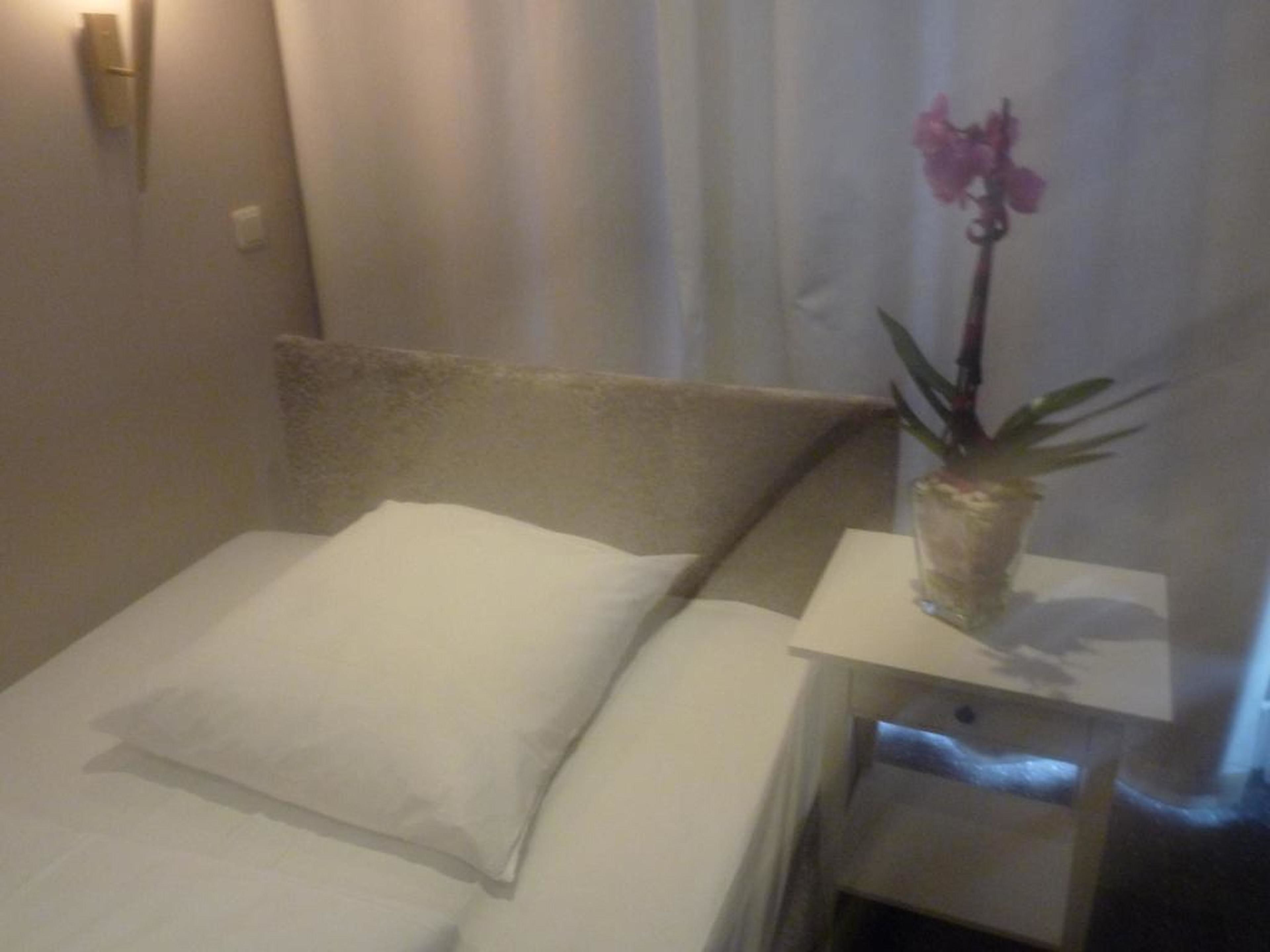 A small, neatly made bed with a white pillow and a single white sheet. There is a small bedside table with a vase containing a single orchid.