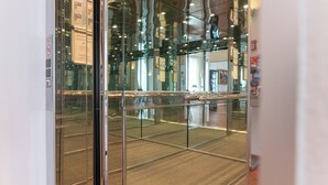 Un ascensor moderno con paneles de vidrio y barandillas de metal, que refleja el interior de un edificio.
