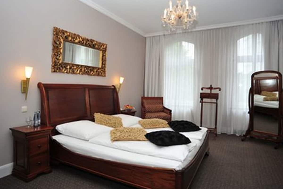 Ein Hotelzimmer mit einem großen Bett, einem hölzernen Kopfteil, einer Kristallleuchtre, einem Spiegel, einem Sessel und einem Nachttisch.
