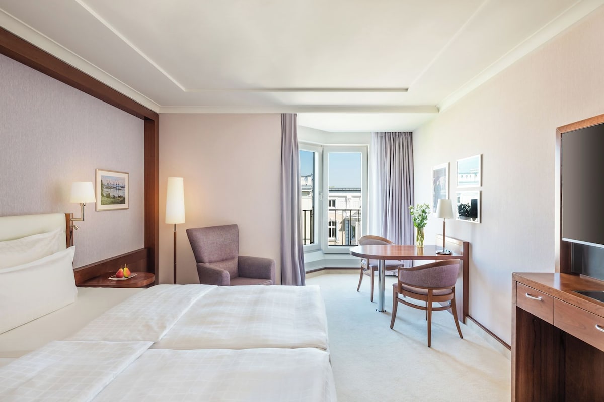 Habitación de hotel con cama, escritorio, silla, televisor y balcón con vistas.