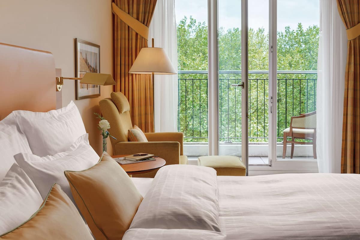 Habitación de hotel con una cama grande, una silla, una mesa y vistas al exterior desde un balcón.