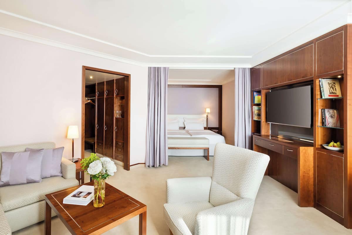 Habitación de hotel con sofá, sillón, mueble de televisión de madera y cama con cabecera.