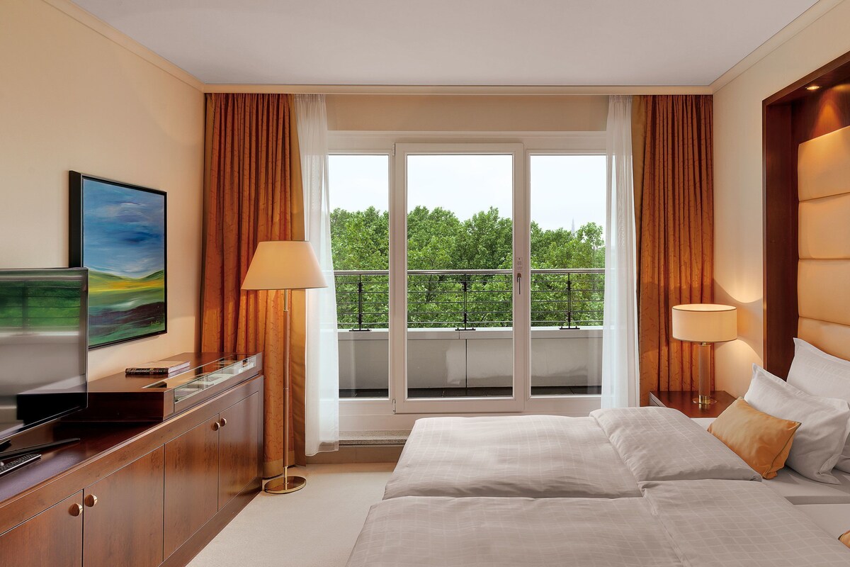 Habitación de hotel con cama, dos lámparas de noche, televisor, escritorio con lámpara y balcón con vista a árboles.