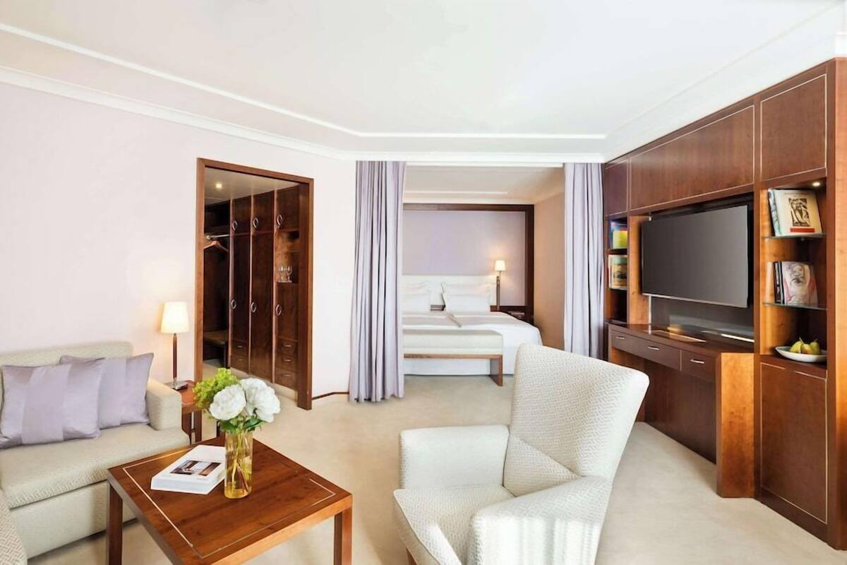 Habitación de hotel con sofá, sillón, mueble bajo de madera para el televisor y cama con cortinas.