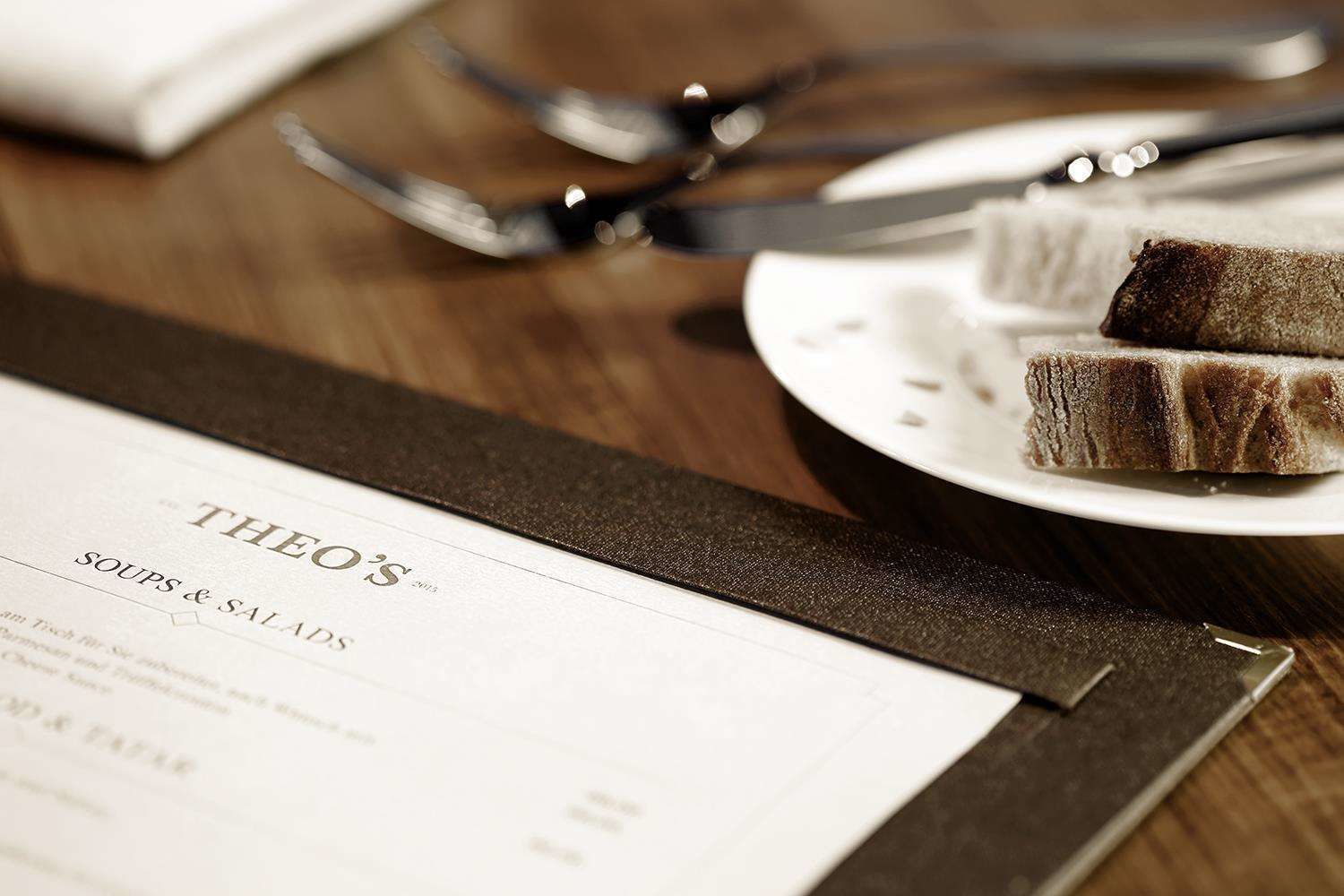 A menu titled "THEO'S SOUPS & SALADS" on a wooden table with a plate of cake and cutlery.