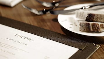 A menu titled "THEO'S SOUPS & SALADS" on a wooden table with a plate of cake and cutlery.