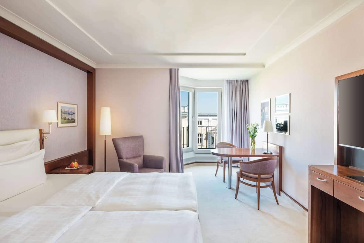 Habitación de hotel con dos camas, un escritorio con silla, televisión y balcón con vistas.