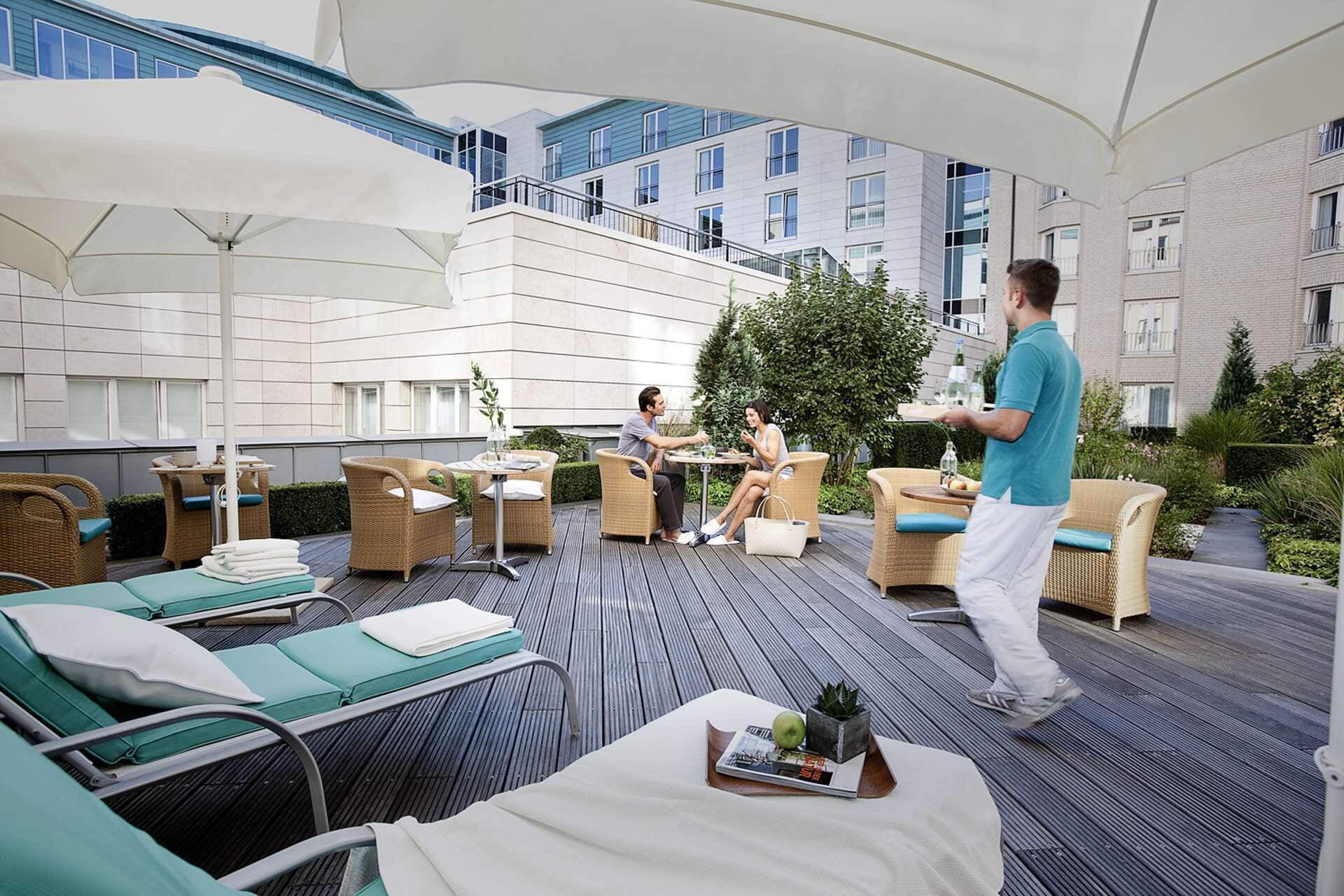 A rooftop patio with wicker furniture, a waiter serving drinks, and a view of the building's exterior.