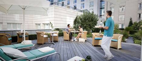 A rooftop patio with wicker furniture, a waiter serving drinks, and a view of the building's exterior.