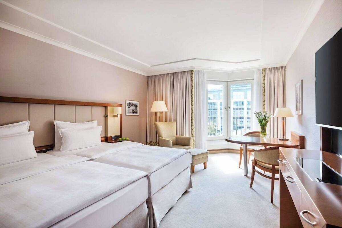 Habitación de hotel con dos camas, un escritorio, una silla, un televisor y un ventanal con cortinas.