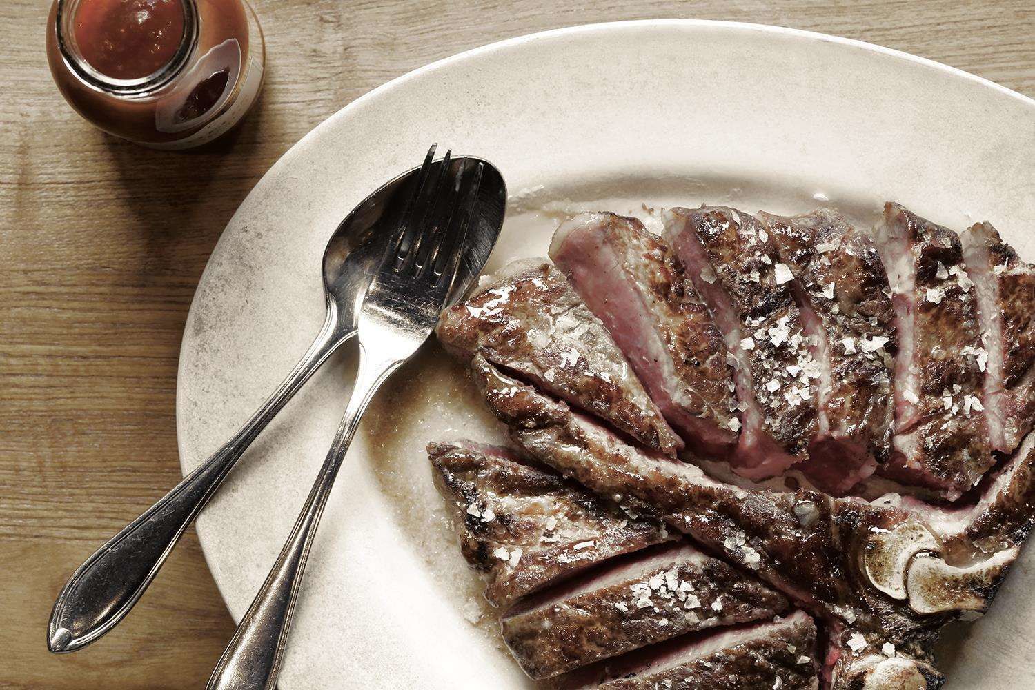 A plate of sliced meat with a fork and knife, and a jar of sauce on a wooden table.