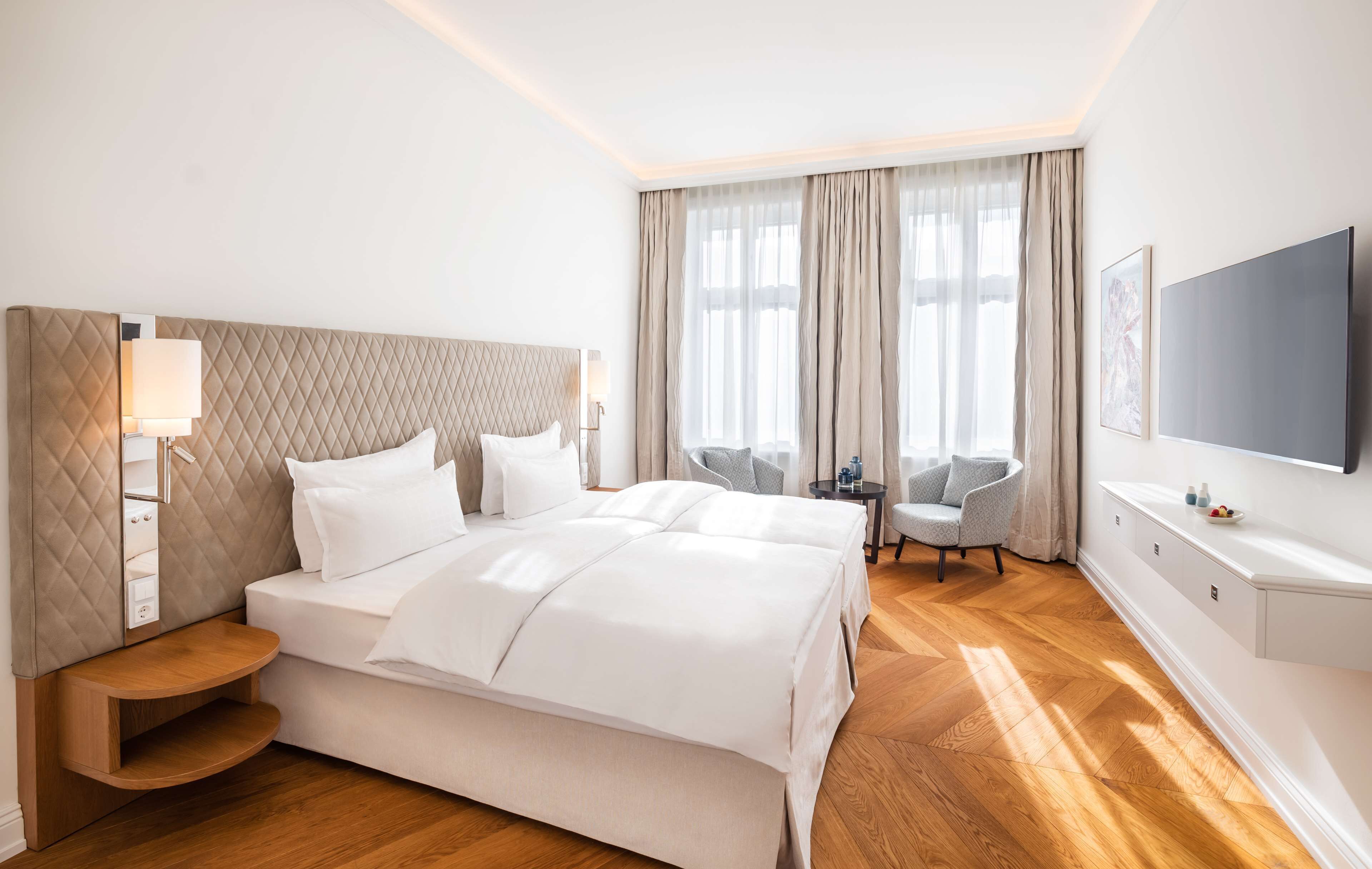 A hotel room with a large bed, bedside tables, a TV, a small seating area, and a large window with curtains.