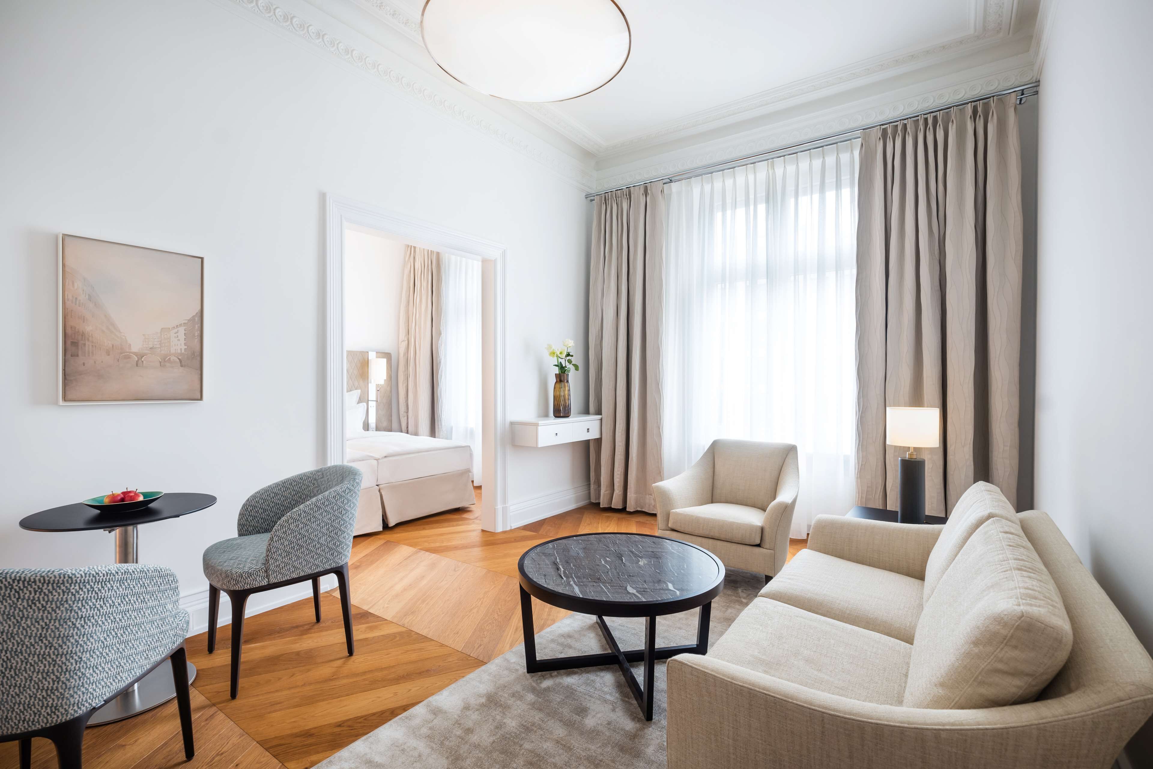 Ein modernes Wohnzimmer mit einer Couch, zwei Sesseln, einem Couchtisch und einem Esstisch. Im Hintergrund steht ein Bett, an der Wand hängt ein Gemälde und auf einem Beistelltisch steht eine Lampe.