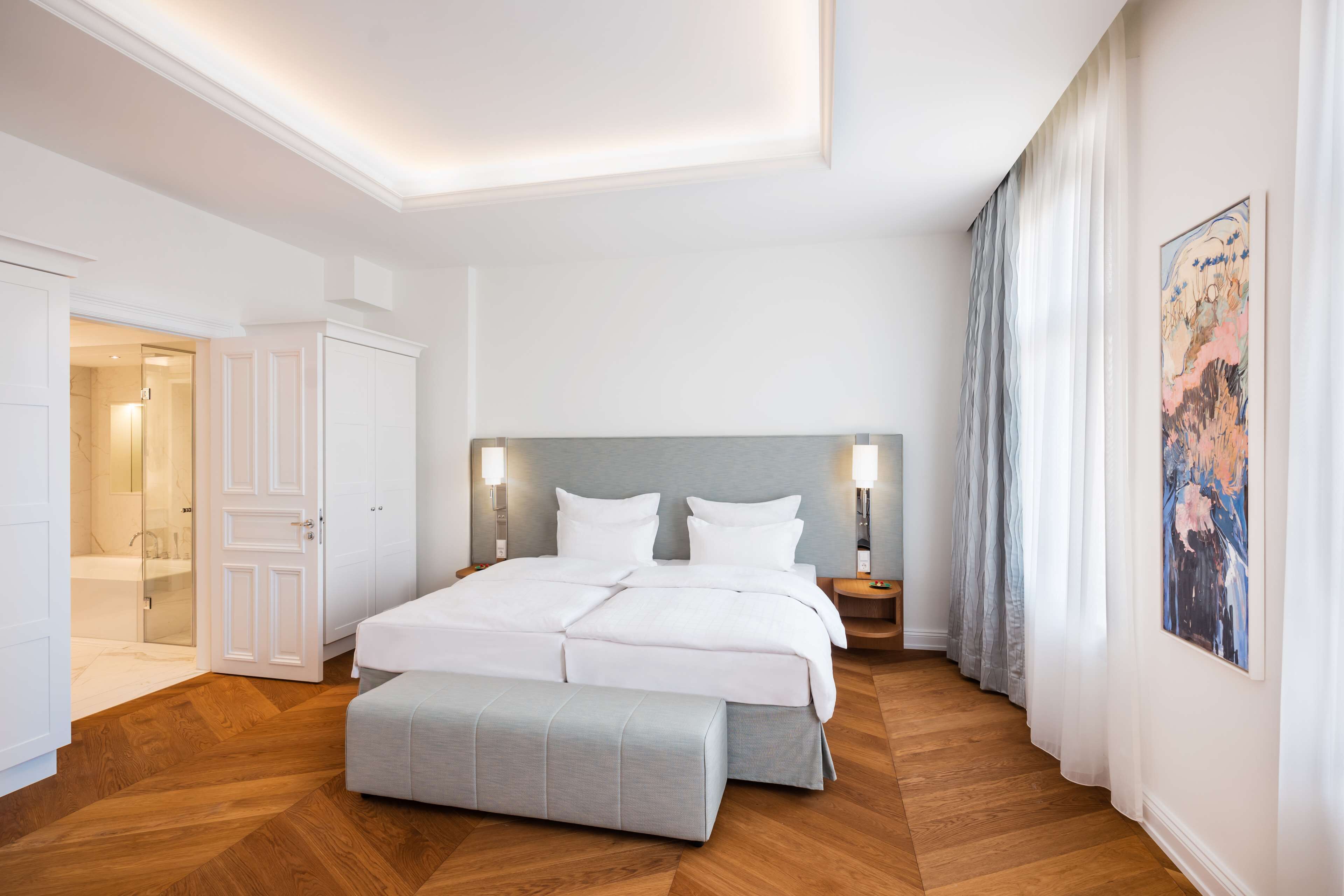 Un dormitorio moderno con una cama grande, una banca, un vestidor y un baño con ducha de vidrio.