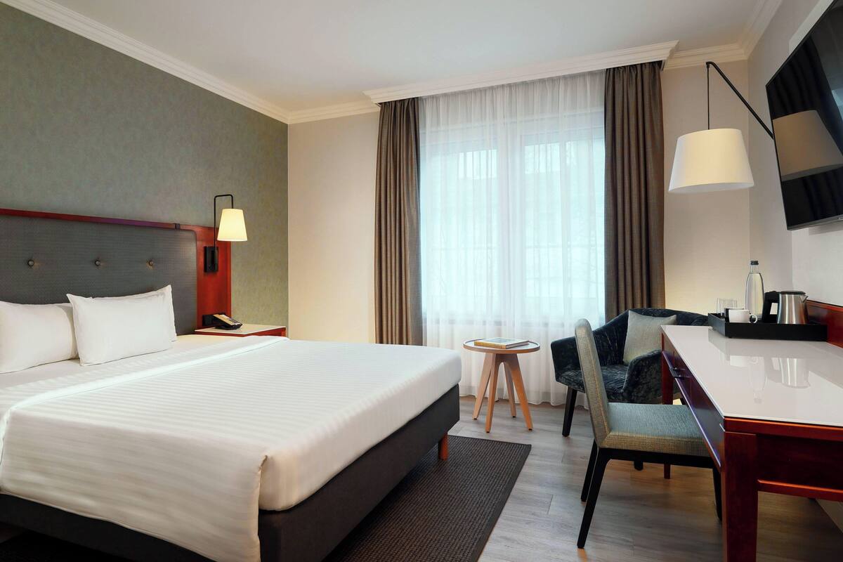 A hotel room with a large bed, a desk with a chair, a TV, and a window with curtains.