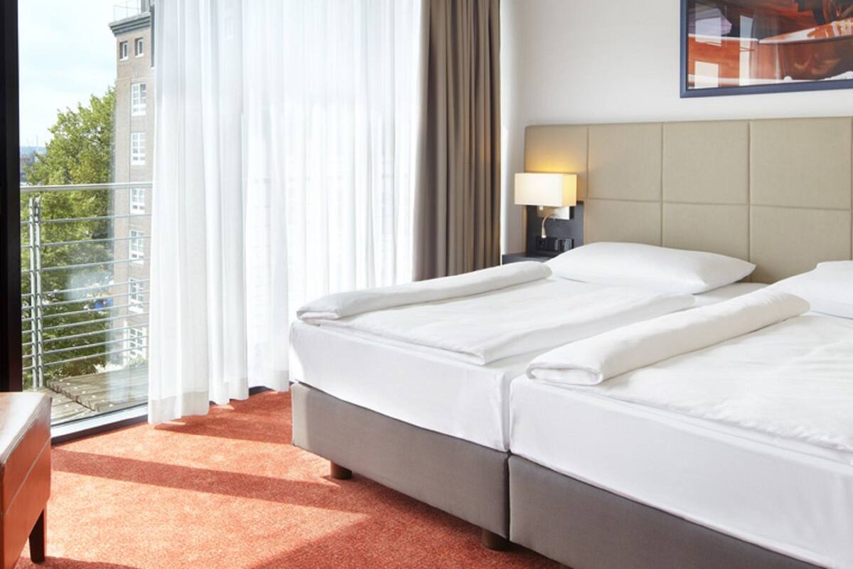 A hotel room with two beds, a balcony with a view of buildings, and a painting on the wall.