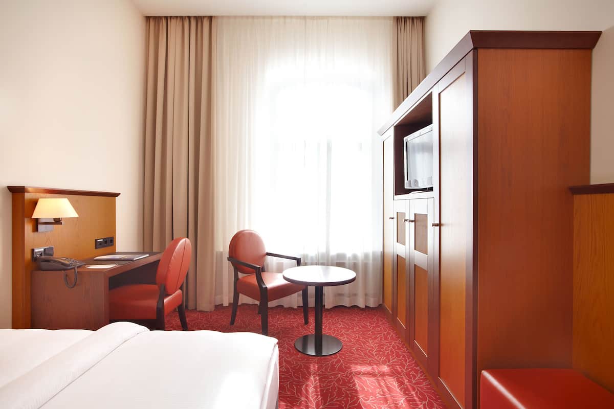 A hotel room with a bed, a desk with a lamp, a chair, a small table, and a TV.