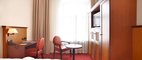 A hotel room with a bed, a desk with a lamp, a chair, a small table, and a TV.