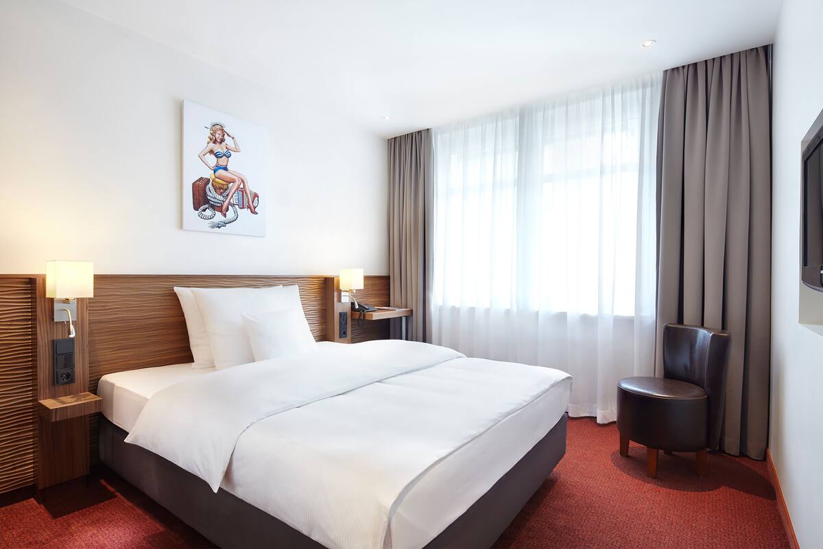 A hotel room with a large bed, a nightstand with a lamp, a chair, a painting on the wall, and a window with curtains.
