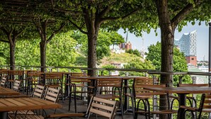 Un café al aire libre con mesas y sillas de madera, rodeado de árboles, con vistas a un paisaje urbano con edificios y una iglesia.