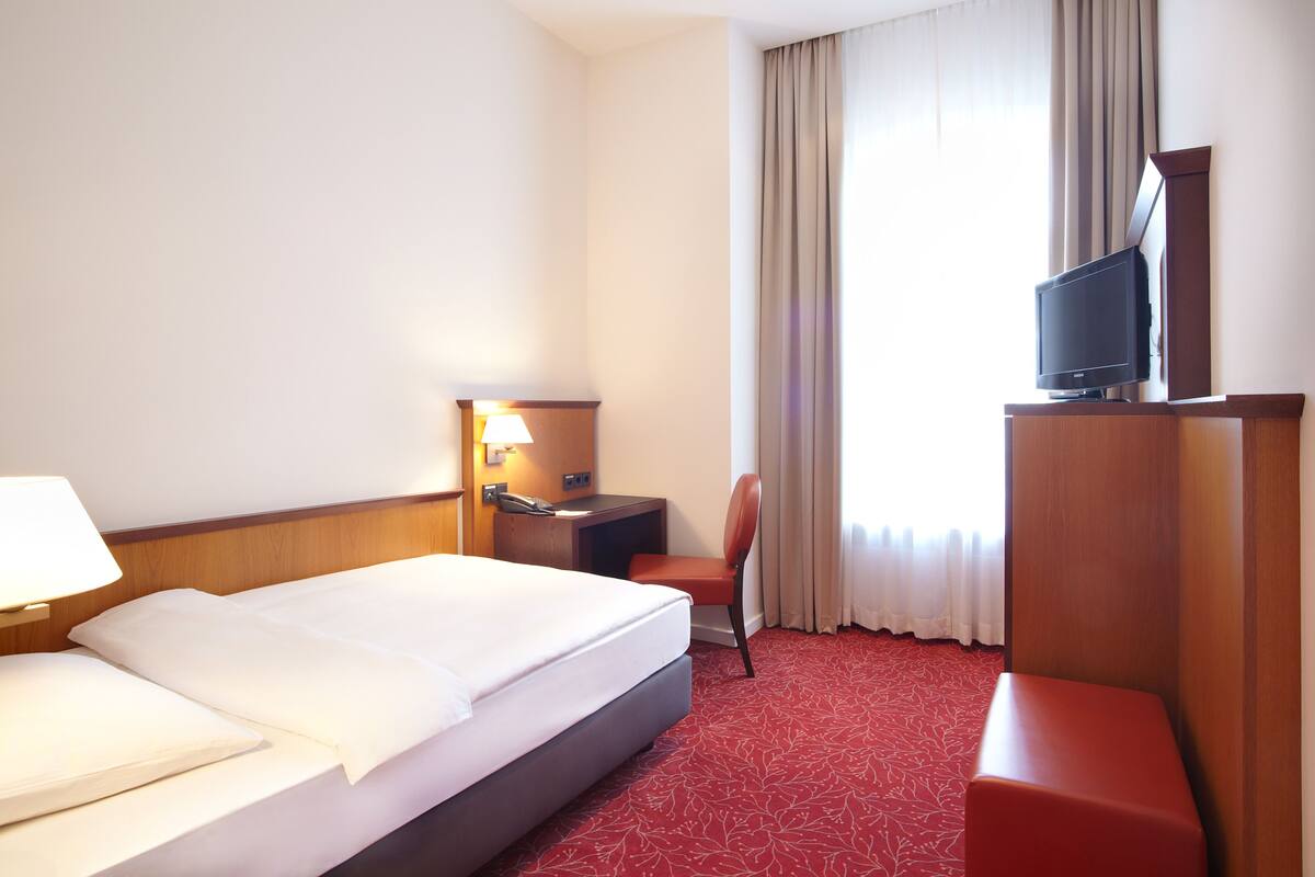A hotel room with a bed, a desk with a lamp, a television on a stand, and a red carpeted floor.