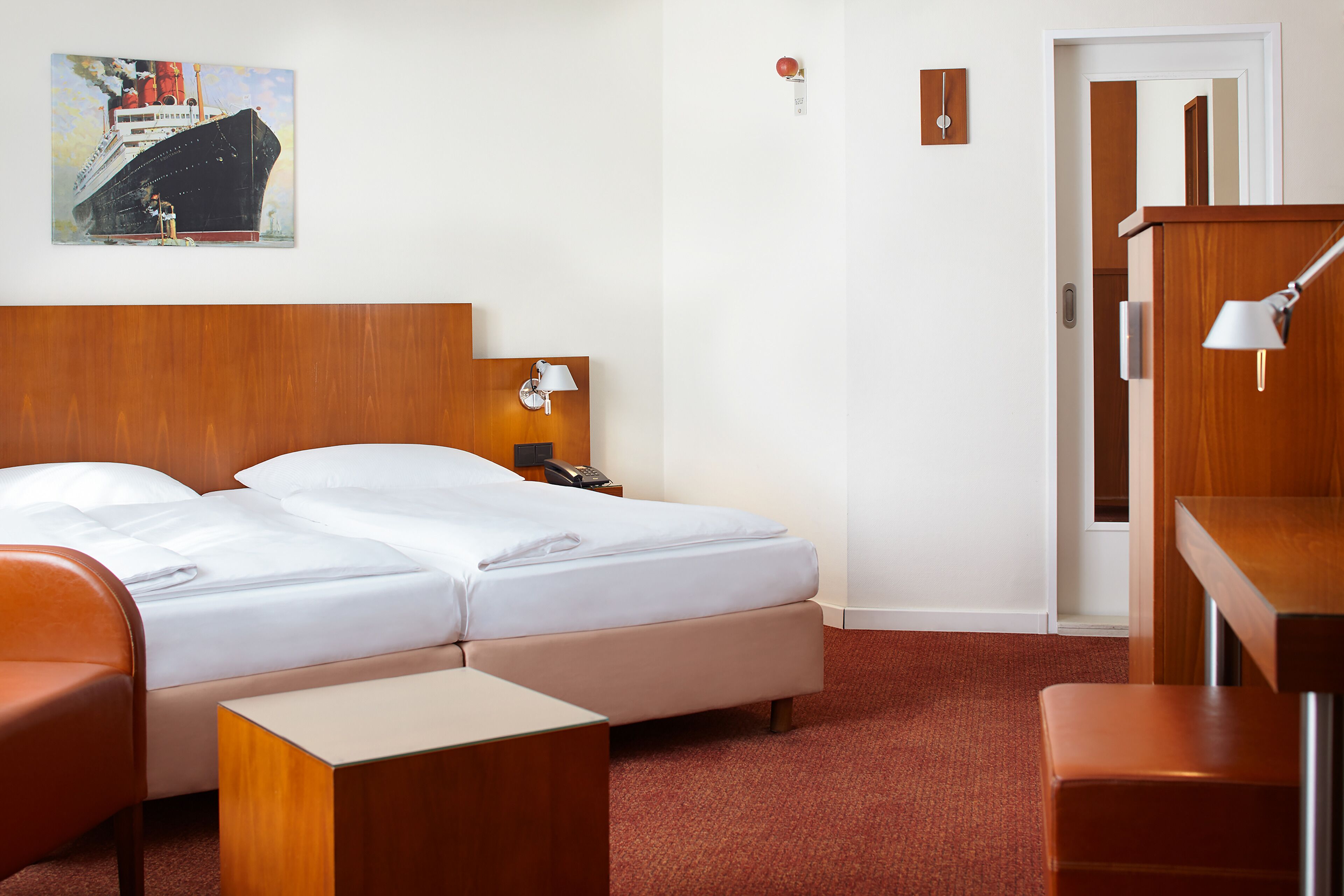 A hotel room with a double bed, a wooden headboard, a desk with a lamp, a chair, and a small table.