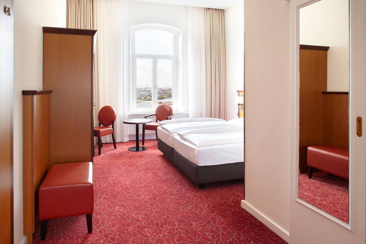 A hotel room with a large bed, a desk, a chair, and a view of the city through a large window.