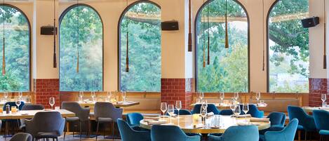Ein Restaurant mit runden Tischen, blauen Stühlen und großen Bogenfenstern mit Blick auf Grünflächen.
