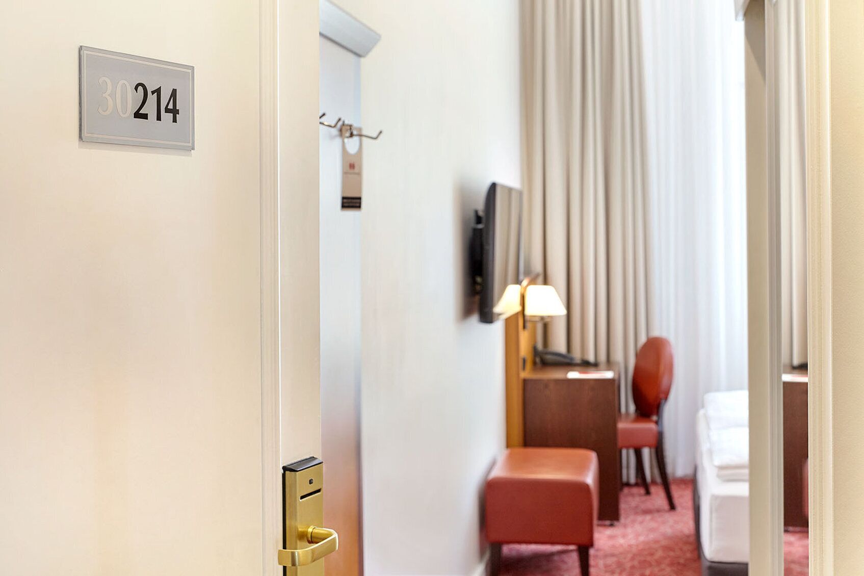 A hotel room with a door labeled "30214", a desk with a lamp, a chair, and a bed.