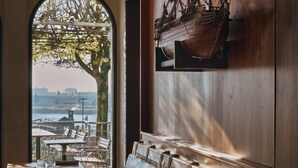 Un restaurante con una larga banqueta de madera, un amplio ventanal arqueado con vistas a un paisaje urbano y un modelo de barco en la pared.