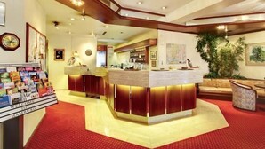 Un espace d’accueil d’hôtel avec un comptoir central, un présentoir de magazines et un sol recouvert d’un tapis rouge.